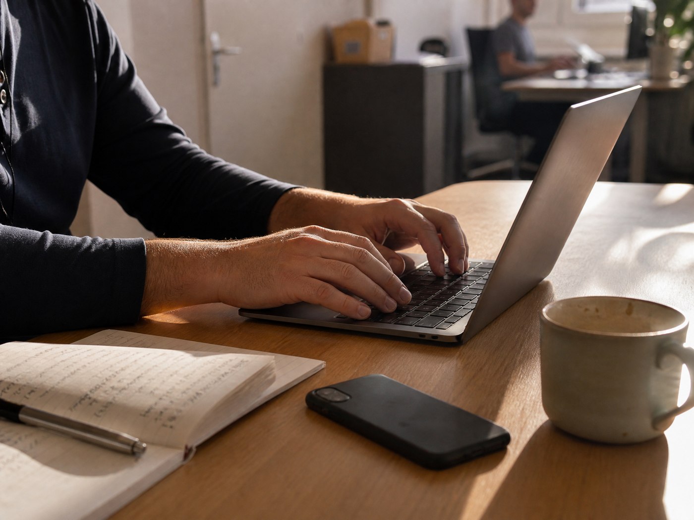 Nahaufnahme: Hände tippen auf einem Laptop, daneben liegt ein privates Smartphone auf dem Bürotisch