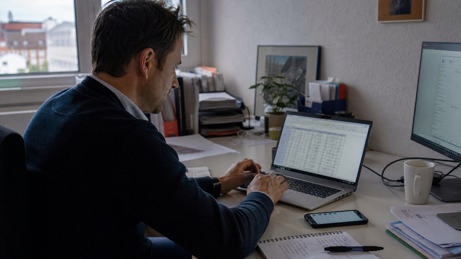 Mitarbeiter arbeitet an einem Laptop mit Firmensoftware, ein Smartphone mit einer privaten Chat-App liegt griffbereit auf dem Schreibtisch daneben